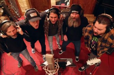 Five musicians wearing headphones sing together around a microphone in a red studio in the IGNITE music video for On The Ropes